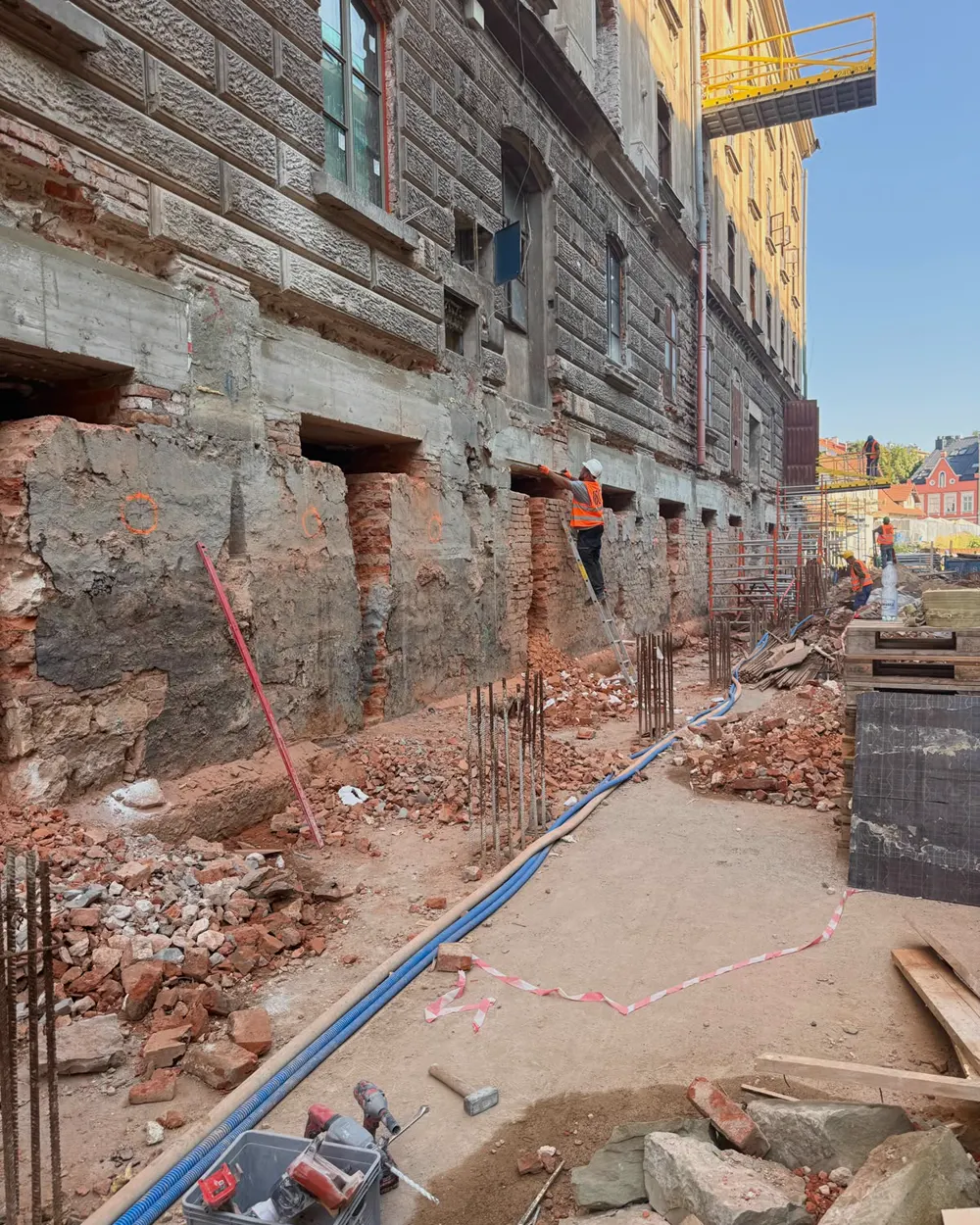 Wykop wzdłuż elewacji zabytkowego budynku — odsłonięty ceglano-kamienny mur fundamentowy z serią wyciętych wnęk i wyprowadzonym zbrojeniem