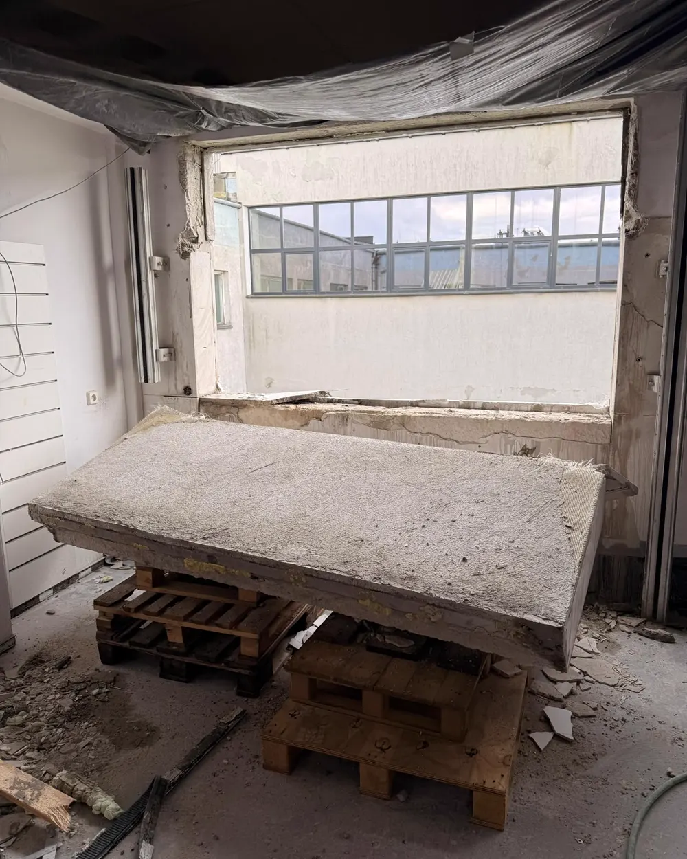 Wycięty blok betonu ułożony na drewnianych paletach we wnętrzu, w tle poszerzony otwór okienny